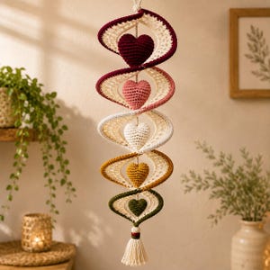 May include: A hanging spiral mobile featuring five crocheted hearts in dark red, light pink, cream, mustard yellow, and dark green. Each heart is centered within a cream-colored crocheted spiral frame, suspended by a cord ending in a cream tassel.