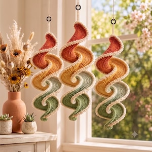 May include: Three hanging spiral decorations crafted from crocheted yarn in a gradient of burnt orange, golden yellow, and sage green. Each spiral features a cream-coloured scalloped edge and is suspended by a thin cord from a black ring.