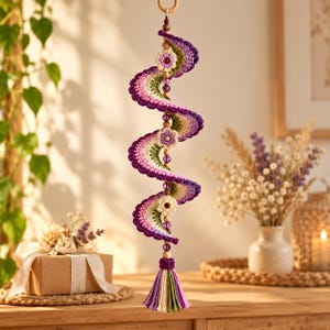 May include: A spiral-shaped crochet hanging ornament featuring layers of purple, green, and cream yarn. The design includes three small crocheted flowers, interspersed with wooden and purple translucent beads, ending in a multi-colored tassel.