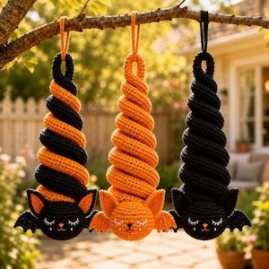 May include: Three hanging crochet ornaments shaped like bats with spiral bodies. The left bat is black and orange, the center is solid orange, and the right is solid black. Each features embroidered closed eyes, small ears, and tiny white fangs.