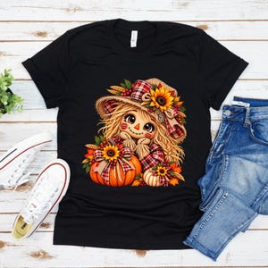 May include: A black t-shirt featuring a graphic illustration of a scarecrow with blonde hair, wearing a plaid hat adorned with a sunflower. The scarecrow is positioned behind two orange pumpkins decorated with sunflowers, plaid ribbons, and autumn leaves.