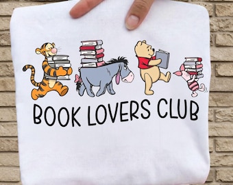 Könnte beinhalten: Ein weißes T-Shirt mit einer Grafik von Tigger, I-Aah, Winnie Puuh und Ferkel, die Bücherstapel tragen. Unter den Figuren steht der Text BOOK LOVERS CLUB in einer fetten, schwarzen, serifenlosen Schriftart auf dem Stoff gedruckt.