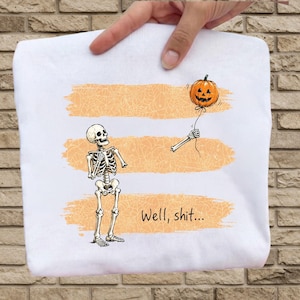 May include: A white t-shirt featuring a graphic of a skeleton looking at its own detached arm holding an orange jack-o'-lantern balloon. The design includes three horizontal orange brushstroke stripes with a spiderweb pattern and the text Well, shit...