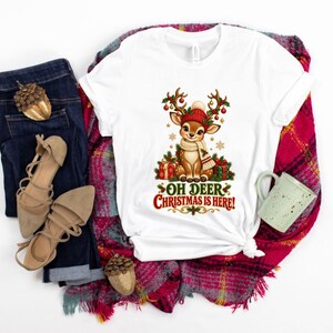 May include: A white short-sleeve t-shirt featuring a graphic of a fawn wearing a red knit hat and scarf, surrounded by Christmas gifts and holly. The text below the fawn reads, "OH DEER CHRISTMAS IS HERE!" The shirt is displayed with dark blue jeans and a plaid scarf.