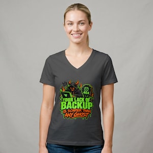 Peut inclure: Un t-shirt gris foncé à col en V avec un motif représentant une silhouette à capuche avec des cornes utilisant un ordinateur portable à côté d'une pierre tombale. Le texte indique : YOUR LACK OF BACKUP IS SCARIER THAN ANY GHOST, en vert et orange.