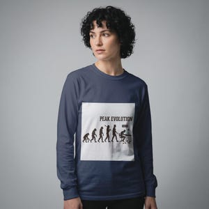 Puede incluir: Una camiseta azul marino de manga larga con un gráfico cuadrado blanco en el pecho. El gráfico muestra una silueta negra de la evolución humana, terminando con una figura sentada en un escritorio con un ordenador y el texto PEAK EVOLUTION.
