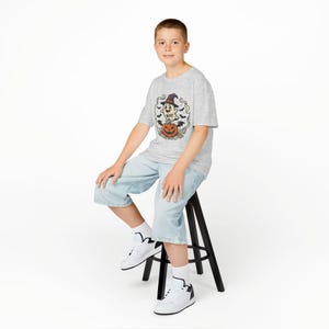May include: A light grey short-sleeved t-shirt featuring a graphic of a smiling ghost wearing a witch hat, sitting on a carved orange pumpkin. The design includes the text "BOO!" and small black bats flying around the central illustration.