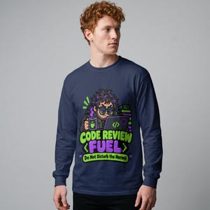 May include: A navy blue long-sleeve shirt featuring a graphic of a person with messy hair and glasses working on a laptop while holding a mug. The text reads CODE REVIEW FUEL and Do Not Disturb the Hermit in green and purple lettering.