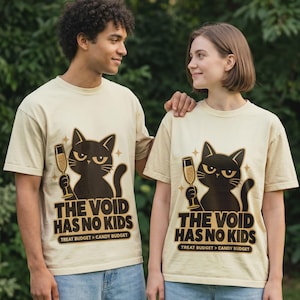 May include: A beige short-sleeve t-shirt featuring a graphic of a black cat holding a champagne flute. The text reads THE VOID HAS NO KIDS and TREAT BUDGET > CANDY BUDGET. The design includes small star accents in a dark brown and gold color palette.