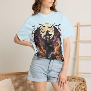 Puede incluir: Una camiseta azul claro con un estampado gráfico de una bruja con túnica y sombrero negros que sostiene un bastón. La escena incluye una gran luna llena, árboles desnudos, murciélagos voladores, fantasmas y calabazas naranjas de fondo.