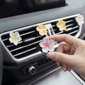 May include: A set of car air freshener clips shaped like cherry blossom flowers in shades of white, pale yellow, and soft pink. Each flower features a detailed centre with yellow or pink stamens, designed to attach to vehicle air conditioning vents.