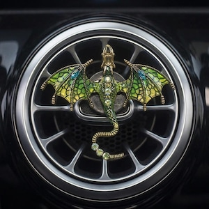 May include: A decorative car air vent clip shaped like a dragon with green and yellow enamel wings, a textured body, and clear crystal accents along the spine and tail. The dragon features blue crystal details on its wings and a gold-tone finish.