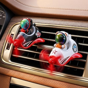 Può includere: Due clip deodoranti per auto a forma di piccoli aerei con piloti orso che indossano caschi iridescenti. Un aereo è nero e l'altro è bianco, entrambi dotati di eliche rosse, grafiche con denti di squalo e decalcomanie a stella sui lati.