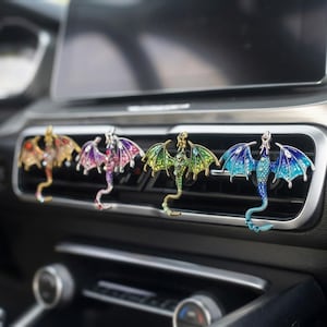 May include: Four metal dragon-shaped car air vent clips are attached to a vehicle dashboard vent. Each dragon features colorful enamel wings and small embedded crystals, with one in red and gold, one in purple and pink, one in green, and one in blue.