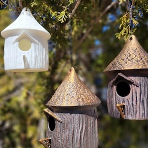 Puede incluir: Tres casas para pájaros colgantes con techos cónicos y orificios de entrada circulares. Una casa es de color blanco sólido con un exterior texturizado similar a la corteza. Dos casas tienen cuerpos marrones oscuros y techos dorados.