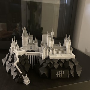 Könnte beinhalten: Ein 3D-gedrucktes Modell einer Burg mit weißen Türmen und Gebäuden auf gezackten schwarzen Felsformationen. Die Struktur umfasst eine Brücke, die zwei Abschnitte verbindet, und eine schwarze Basisplatte mit den Buchstaben HP in stilisierter Schrift.
