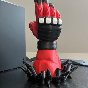 May include: A red and black sculpted hand figurine featuring a tactical glove design with white knuckle guards. The base of the wrist is surrounded by a circular arrangement of black bullet-shaped decorative elements, creating a detailed display piece.
