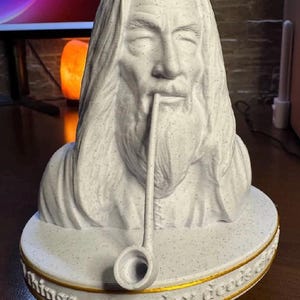 Puede incluir: Un busto blanco moteado de una figura barbuda con cabello largo, sosteniendo una pipa larga en la boca. La figura se asienta sobre una base circular con un borde pintado en oro y el texto en relieve: "things, everyday deeds do."