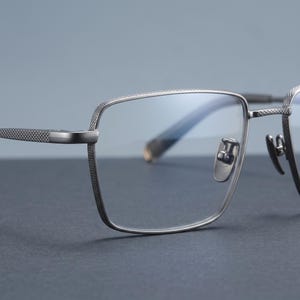 Blue light blocking clear demo lenses on gunmetal grey titanium eyeglasses, a macro of the square optical frame showing the faint anti-glare blue sheen across the lens for office wear.