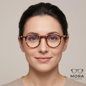 May include: Tortoiseshell-patterned spectacles with a round frame, a thin metal bridge, and silver-toned rivets at the hinges. The frame features a classic mottled brown and amber design. The logo MORA EYEWEAR is printed in the bottom right corner.