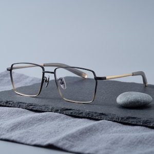 Black and gold titanium eyeglasses styled still life on natural linen, a two-tone square optical frame for men showcased as minimalist business eyewear for men.