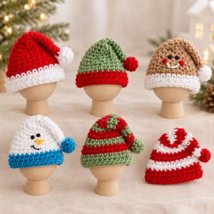 May include: Six miniature crochet hats displayed on wooden stands. The collection includes a red and white Santa hat, a green and red elf hat, a tan gingerbread-style hat, a white snowman hat with a blue brim, a green and red striped hat, and a red and white striped hat.