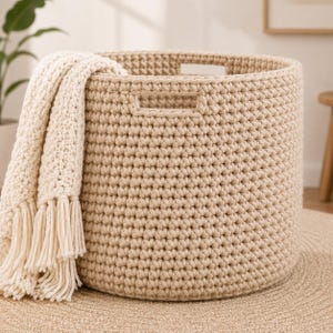 Op de afbeelding: Een ronde, beige, gehaakte opbergmand met geïntegreerde handgrepen aan de zijkant. Een dikke, crèmekleurige, grofgebreide deken met lange, geknoopte franjes is over de linkerkant van de mand gedrapeerd en bedekt gedeeltelijk de buitenwand.