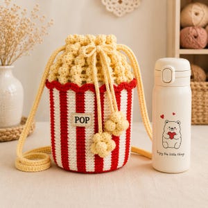 May include: A crochet drawstring bag designed to look like a popcorn bucket with red and white vertical stripes, a yellow textured top, and a rectangular patch labeled POP. Beside it stands a cream-colored insulated bottle featuring a bear graphic.