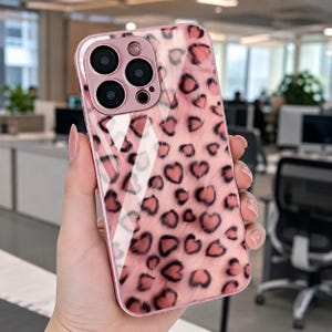 May include: A glossy smartphone case featuring a pink background with a repeating pattern of dark-outlined heart-shaped spots. The case has a metallic pink camera module cutout with three circular lens openings and a small flash aperture.