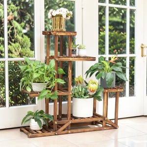 Puede incluir: Un soporte para plantas de varios niveles hecho de madera marrón oscura con un diseño de listones, que cuenta con varios estantes para exhibir plantas en macetas. El soporte sostiene varios follajes verdes, plantas con flores en macetas blancas y grises.