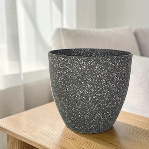 Puede incluir: Una maceta de color gris oscuro con un patrón de motas blancas en toda su superficie exterior. El recipiente presenta una abertura circular ancha en la parte superior y se estrecha ligeramente hacia la base, con una textura lisa y uniforme.