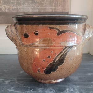 Könnte beinhalten: Ein Keramiktopf mit zwei seitlichen Griffen, einer gesprenkelten beigen Grundfarbe, einem dunkelbraunen Rand und abstrakten orangefarbenen und schwarzen bemalten Formen auf der Außenseite. Das Gefäß hat einen runden Körper und eine glatte Oberfläche.