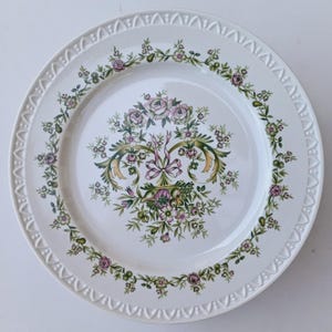 Op de afbeelding: Een wit keramieken bord met een centraal bloemmotief van roze rozen, groen blad en een decoratieve strik. De rand is voorzien van een reliëf met een herhalend boogpatroon en een bloemenslinger omcirkelt het binnengedeelte van het bord.