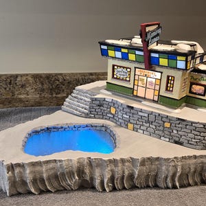 May include: A miniature bowling alley building titled Memory Lanes with a stone foundation, a blue illuminated pond, and a staircase. The structure features a Pro Shop, a Spare-time Lounge, and a Basement Bowl sign, all set on a textured base.