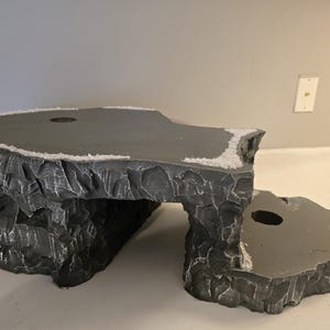 May include: A two-tiered gray rock-like structure with a textured, craggy exterior and smooth, flat top surfaces. Each level features a circular opening. The edges of the upper platform are accented with a thin layer of white, granular material.