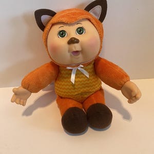 May include: A plush doll with green eyes and rosy cheeks wearing an orange fox costume. The costume features a hood with dark brown ears, a yellow and orange chevron patterned torso, a white ribbon bow at the neck, and dark brown feet.