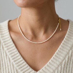 Könnte beinhalten: Eine zarte Halskette aus kleinen, gleichmäßigen cremeweißen Perlen in einer einreihigen Anordnung. Die Kette verfügt über eine goldfarbene Metallverlängerung an einer Seite, die mit einer kleinen Perle endet, sowie einen sicheren Karabinerverschluss.