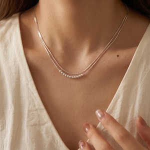 May include: A double-strand silver necklace featuring a delicate chain design. The lower strand is adorned with a series of small, polished silver beads arranged in a horizontal row, creating a subtle and refined look against the neckline.