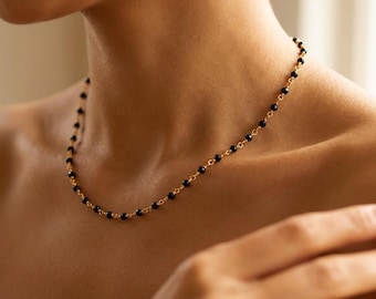May include: A delicate gold chain necklace featuring small, round, polished black beads spaced evenly along the entire length of the chain. The necklace rests against the neck, showcasing a simple link design with dark, spherical accents.