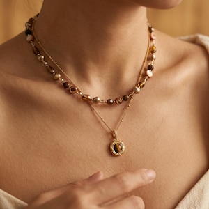 Puede incluir: Un conjunto de collares superpuestos que presenta una hebra más corta de cuentas irregulares en tonos marrón y crema, y una cadena dorada más larga con un colgante ovalado central. El colgante contiene una piedra ojo de tigre rodeada de cristales.