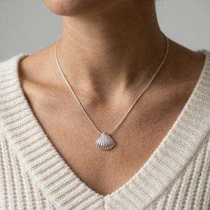 May include: A silver-toned necklace featuring a small, textured seashell pendant suspended from a thin, smooth chain. The pendant has a fan-like shape with vertical ridges, reflecting light across its metallic surface. The chain rests against the neck.