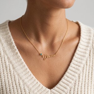 Peut inclure: Un collier doré composé d'une chaîne fine et d'un pendentif nominatif en écriture cursive portant le nom Danica. Le collier est agrémenté d'une petite pierre précieuse ronde de couleur sarcelle placée juste à côté du pendentif sur la chaîne.