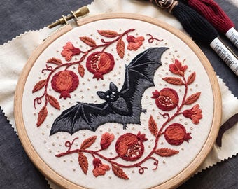 May include: A circular wooden embroidery hoop containing a fabric panel with a hand-stitched design of a black bat centred among red pomegranate fruits, leaves, and small white French knots. Two skeins of Airo embroidery floss are included.
