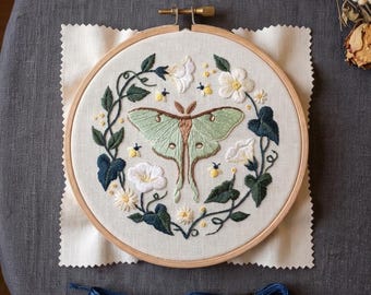 May include: A circular wooden embroidery hoop containing a fabric panel with a hand-stitched design of a pale green luna moth surrounded by white flowers, dark green leaves, and small yellow bees. Two skeins of Airo embroidery floss are below.