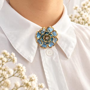 Puede incluir: Un broche dorado con forma de flor de varias capas con pétalos azules translúcidos y un pequeño cristal transparente en el centro, prendido en el cuello de una camisa blanca abotonada con un botón de plástico blanco visible.