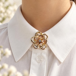 May include: A gold-toned flower brooch featuring multiple clear, faceted crystals arranged in a circular petal pattern. The brooch is secured to the centre of a white collared shirt, positioned just below the neckline, with a single clear button visible.