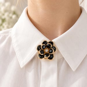 Könnte beinhalten: Eine goldfarbene Brosche in Blumenform mit geschichteten schwarzen, kreisförmigen Blütenblättern und einem einzelnen klaren Kristall in der Mitte. Die Brosche ist am Kragen eines weißen Hemdes befestigt und hält den Stoff am Ausschnitt zusammen.
