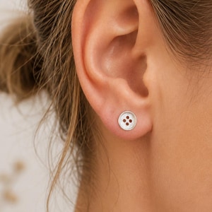 May include: A small, circular silver stud earring shaped like a four-hole sewing button. The earring features a textured, matte metallic finish with a slightly raised outer rim and four distinct circular indentations in the center of the piece.