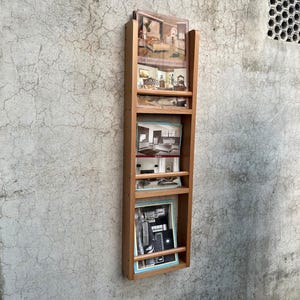 May include: A vertical light brown wooden wall-mounted rack featuring three open shelves. Each shelf holds printed brochures or catalogs displaying interior furniture designs. The rack has a simple frame construction with horizontal support bars.