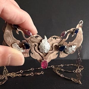 May include: A necklace featuring a wide, textured copper-toned metal centrepiece with organic, leaf-like cutouts. The piece is adorned with small, faceted teardrop-shaped gemstones in deep blue and pink hues, and a delicate, intricate metal chain.